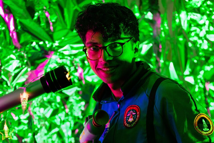 Smiling man wearing glasses in half shadows, with a telescope next to him, standing in front of lush greenery.