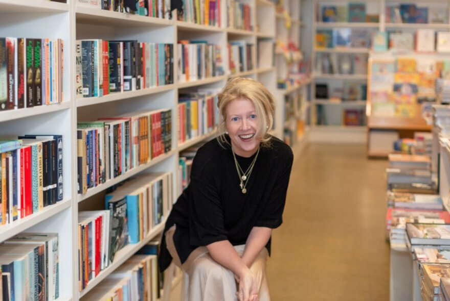 Woman smiling, seated on a chair beside shelves  and stacks of books.