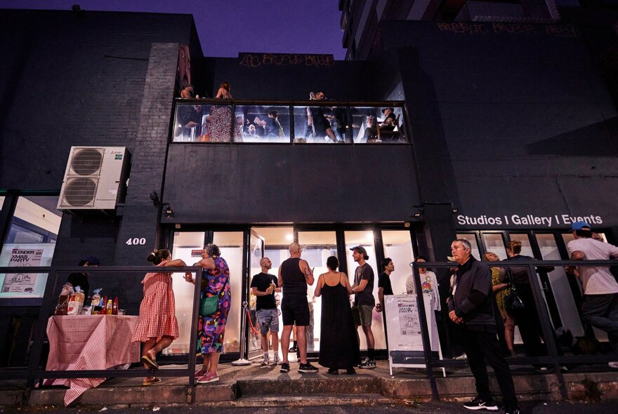 People standing in a laneway at night with drinks and illuminated studio in the background.