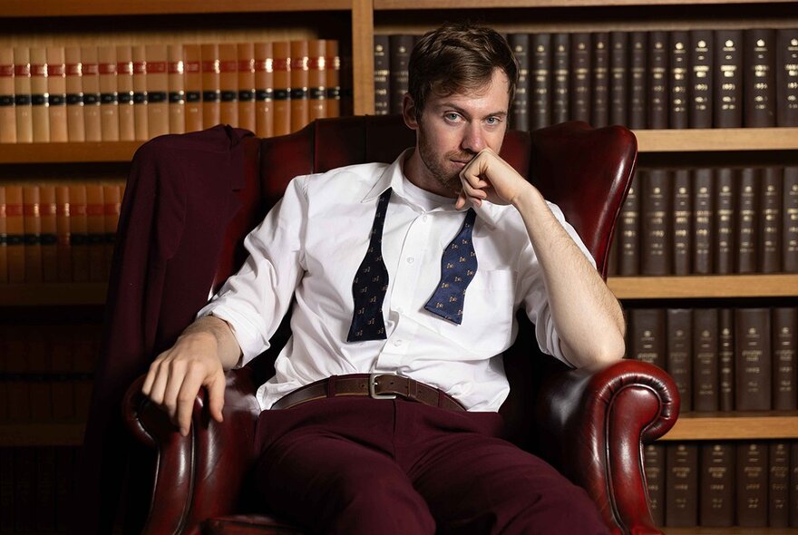 A man reclining in a red leather armchair with rows of leather-bound books behind him.