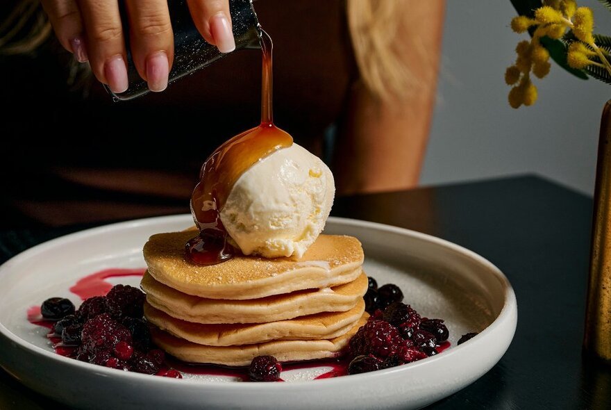 A plate of pancakes with a scoop of vanilla icecream and berries; a woman pouring caramel sauce over it all.