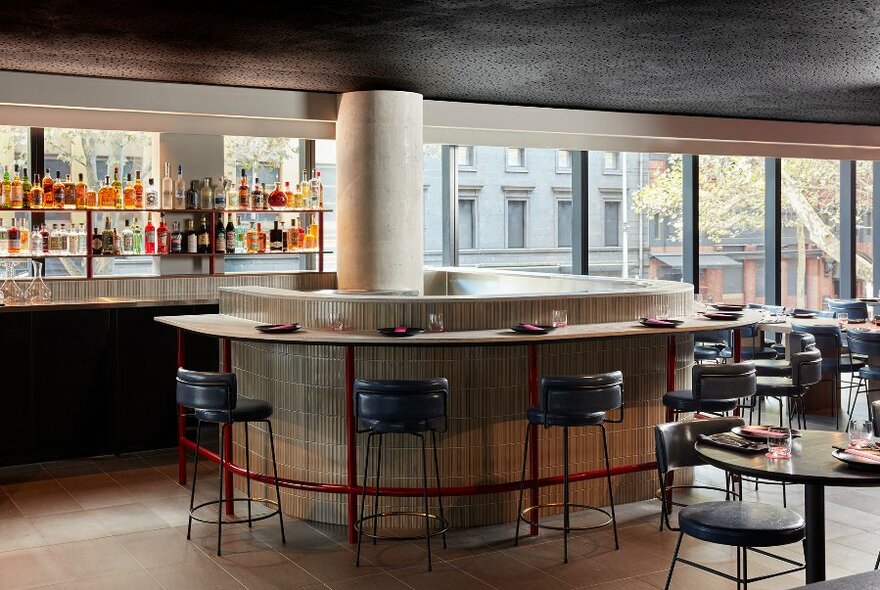 Interior of an empty restaurant with a bar against one wall, large windows overlooking the street, and tables and chairs set for food service.