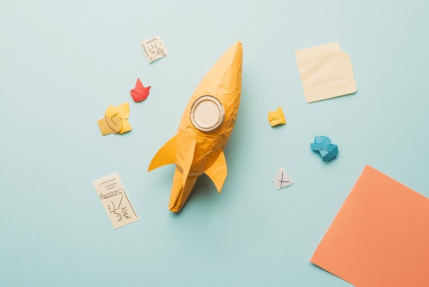 A small three dimensional rocket crafted from yellow paper, with small bits of coloured paper alongside it.