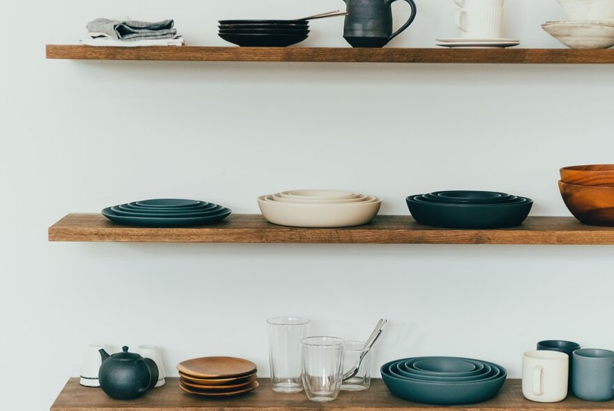 Three wooden shelves of artfully arranged kitchen and ceramic products against a white wall.
