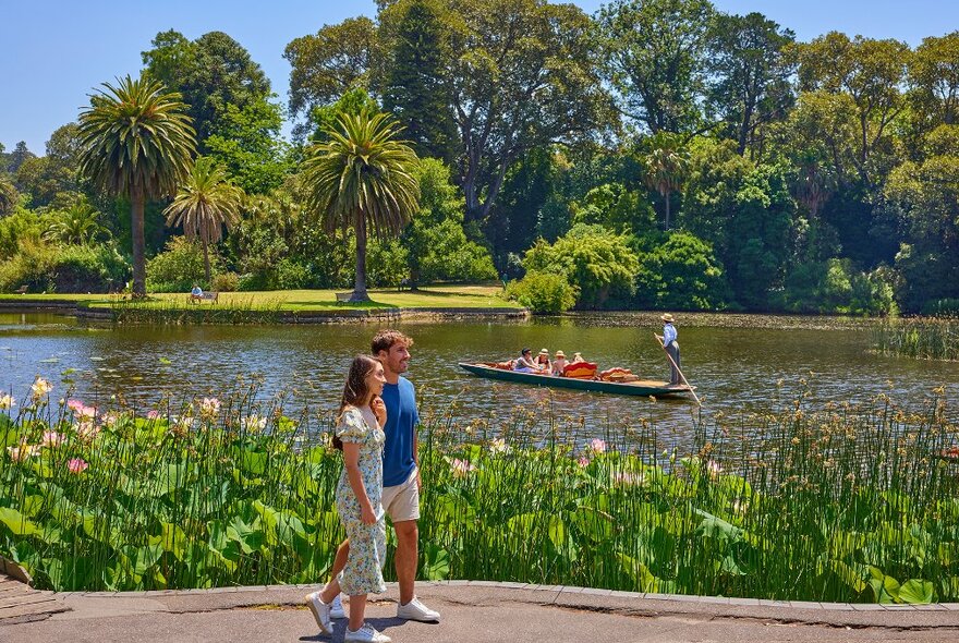 A couple walking beside a lake as a man paddles a punting boat.