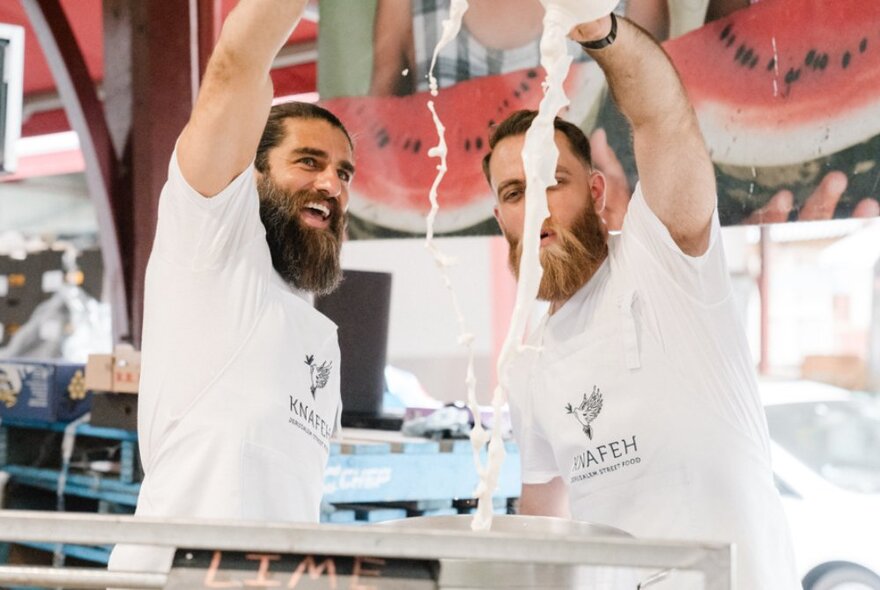 Two bearded men pouring liquid from high in a fruit market with pictures of watermelon behind them.