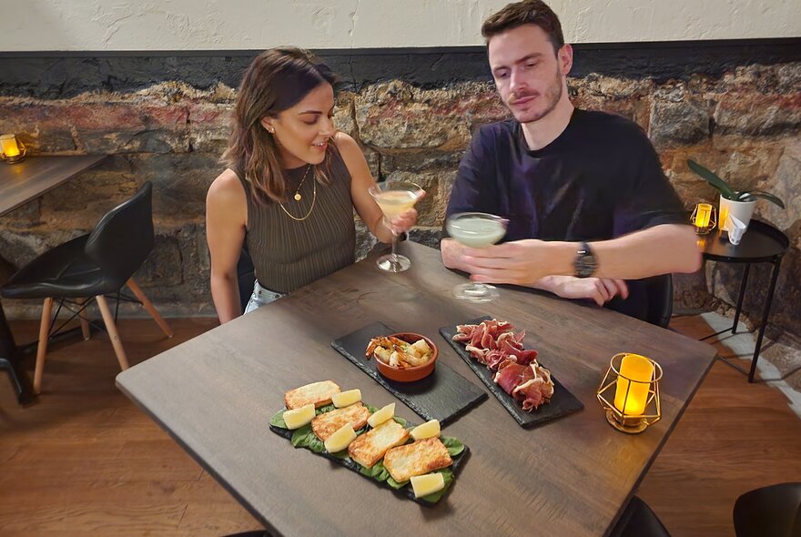 A couple as a table drinking cocktails with antipasto platters of food before them, inside Life's Too Short Bar.