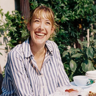 Writer, cook and artist Ella Mittas smiling warmly while sitting at an outdoor table spread with plates of food. 