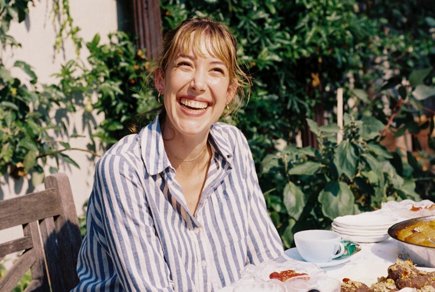 Writer, cook and artist Ella Mittas smiling warmly while sitting at an outdoor table spread with plates of food. 