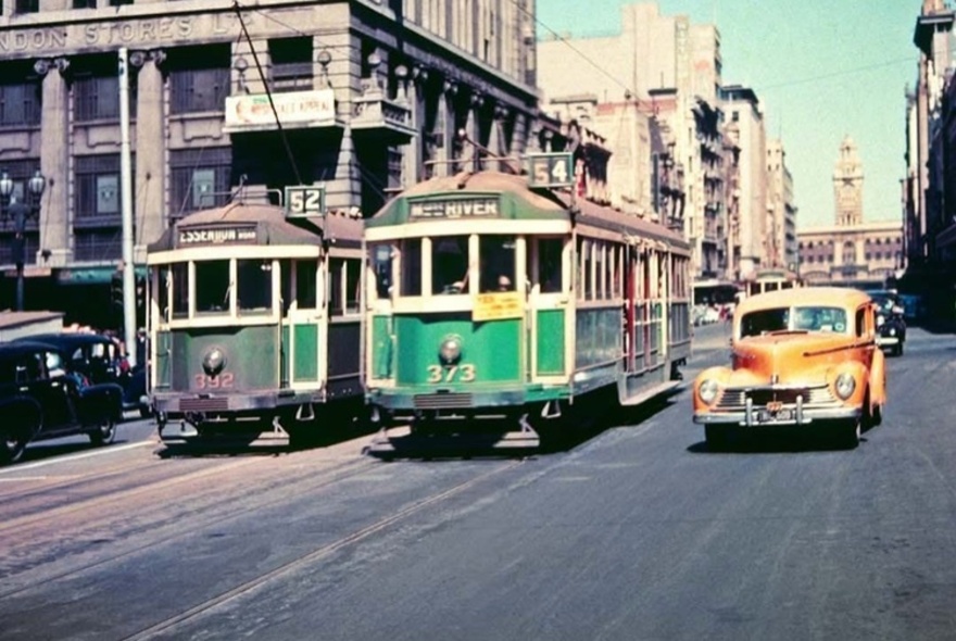 An old colour photograph of two vintage green and yellow W class electric trams travelling on a city street in Melbourne next to a vintage yellow car from the 1950s.