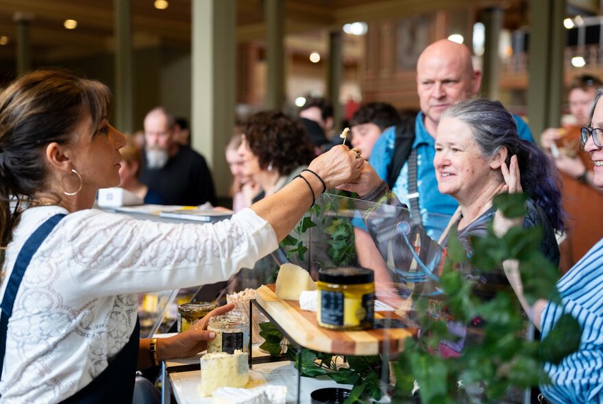 People sampling cheese at a food stall in a market setting.