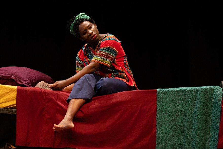 A woman on stage playing reknowned jazz singer and civil rights activist, Nina Simone, as she sits on a simple bed.