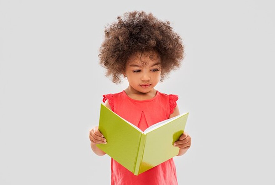 A preschool aged child with curly hair wearing a red dress, holding open a book and looking at it.