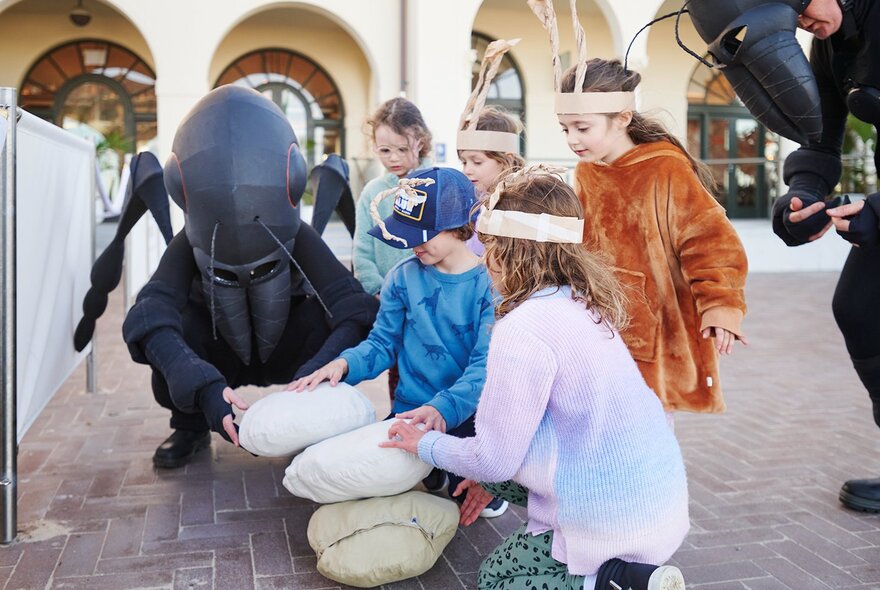 Performers dressed in black ant costumes, interacting with children in stacking giant breadcrumbs, in an outdoor public space.
