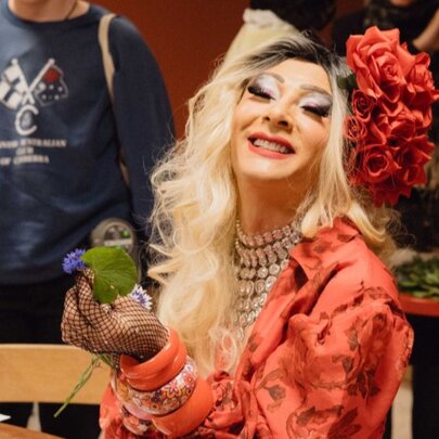 A medium-shot of a smiling person in drag with long blonde wavy hair and a large red floral headpiece, wearing a red patterned jacket, chunky orange bracelets, and a layered necklace.