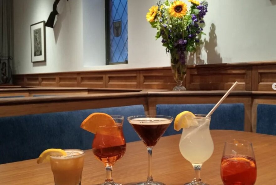 Row of cocktails on a wooden table with blue banquette seating and a vase of sunflowers.