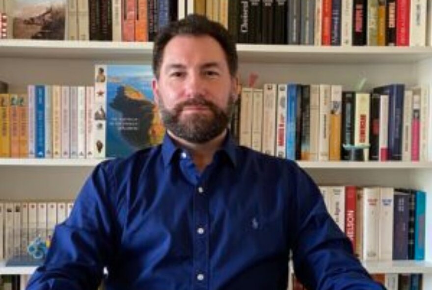 Historian Patrick Llewellyn, a middle-aged man  wearing a blue shirt and looking directly at the viewer, with a bookcase filled with books behind him.