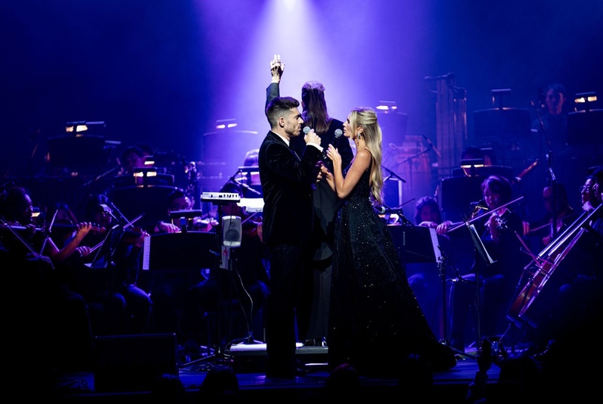 Two singers on stage dressed in formal attire in front of a symphony orchestra with a conductor in the centre.