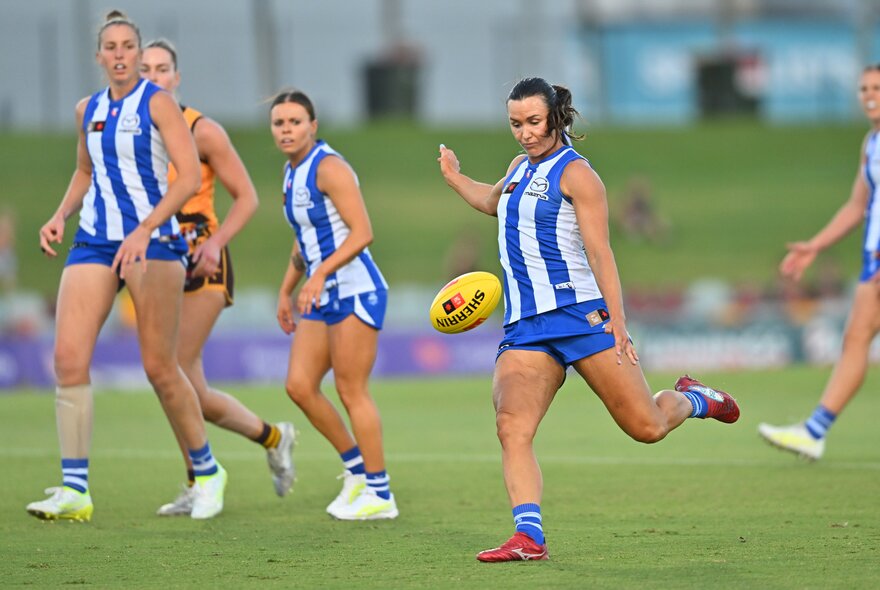 A North Melbourne AFLW player running on the field with the football in her hands about to kick it, with her teammates surrounding her.