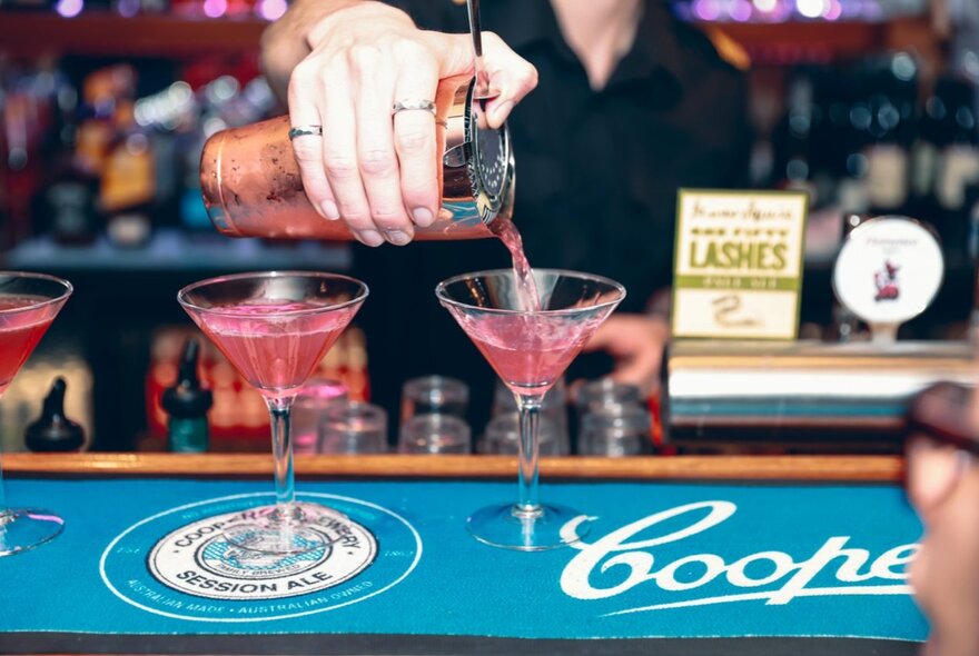 A bartender pouring two cocktails into glasses on a bar counter.