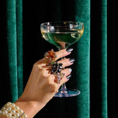 A hand with large rings on its fingers holding up a glass of sparkling wine against a dark green velvet curtain.