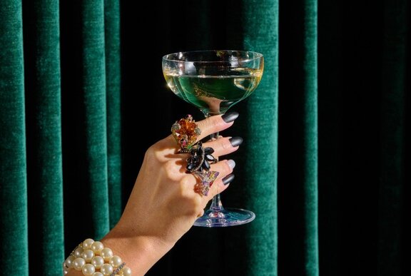 A hand with large rings on its fingers holding up a glass of sparkling wine against a dark green velvet curtain.