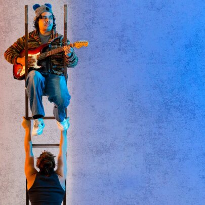 A performer sits at the top of a tall ladder, playing an electric guitar while below them, another performer stands facing away from the camera, holding the ladder steady with uplifted arms.