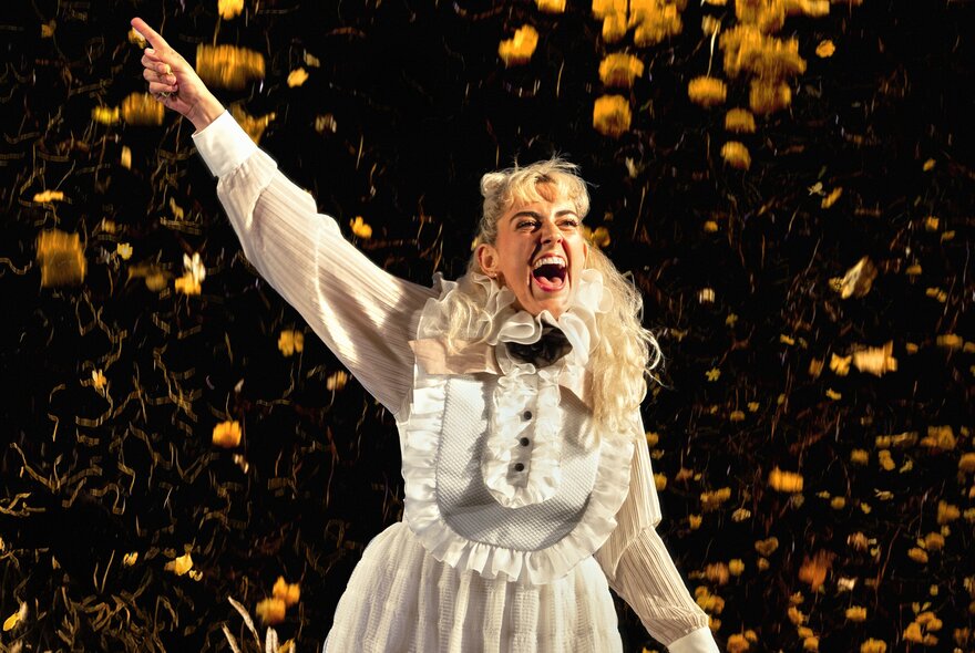 A still image from the musical My Brilliant Career, showing a woman in 1890s costume with her hand in the air and singing while yellow confetti falls around her. 