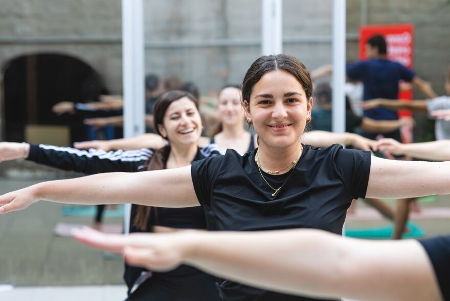 A smiling woman doing yoga in a group setting, others also smiling behind her. 