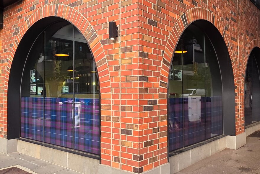 The corner of a red brick building with arched windows with Scottish tartan on the lower half of each window.