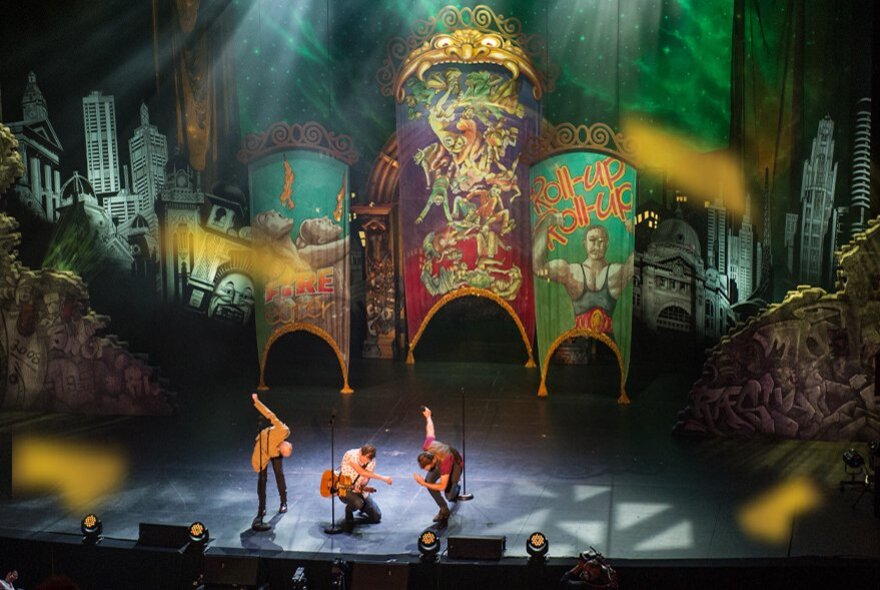 Three comedians on stage in various poses with an elaborately decorated backdrop of various colours.