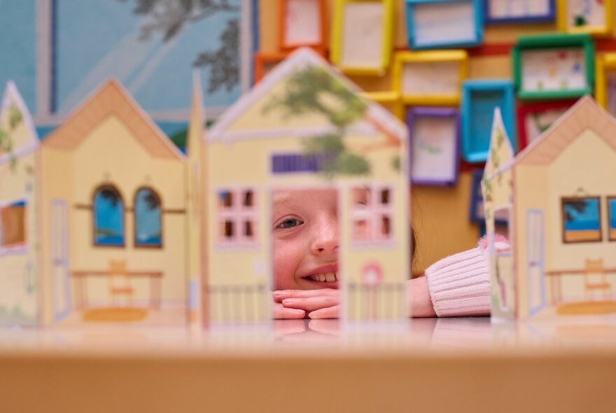 A child's face peeking through a set of interlocking cardboard cut-outs resembling small houses in a row.