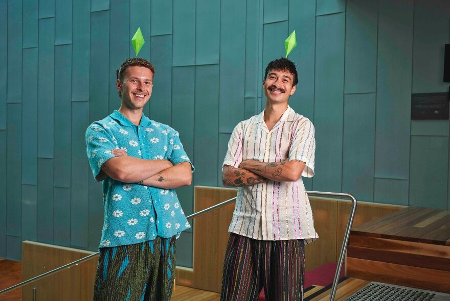 Design duo Josh & Matt Design wearing summery short-sleeved shirts in blue and stripes, with triangles on their heads, standing with arms folded.