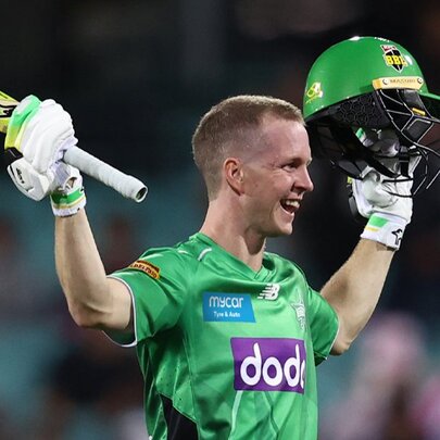 A smiling male cricketer in a bright green Melbourne Stars jersey celebrates a milestone by raising his cricket bat in his right hand and holding up his green helmet in his left hand.