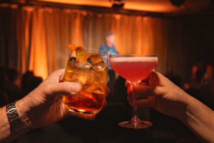 Two hands clinking cocktails in a room with a small stage. 