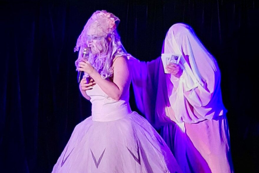 Two performers on stage in a purple light, one singing and the other with a cloak over their head looking like a ghost. 