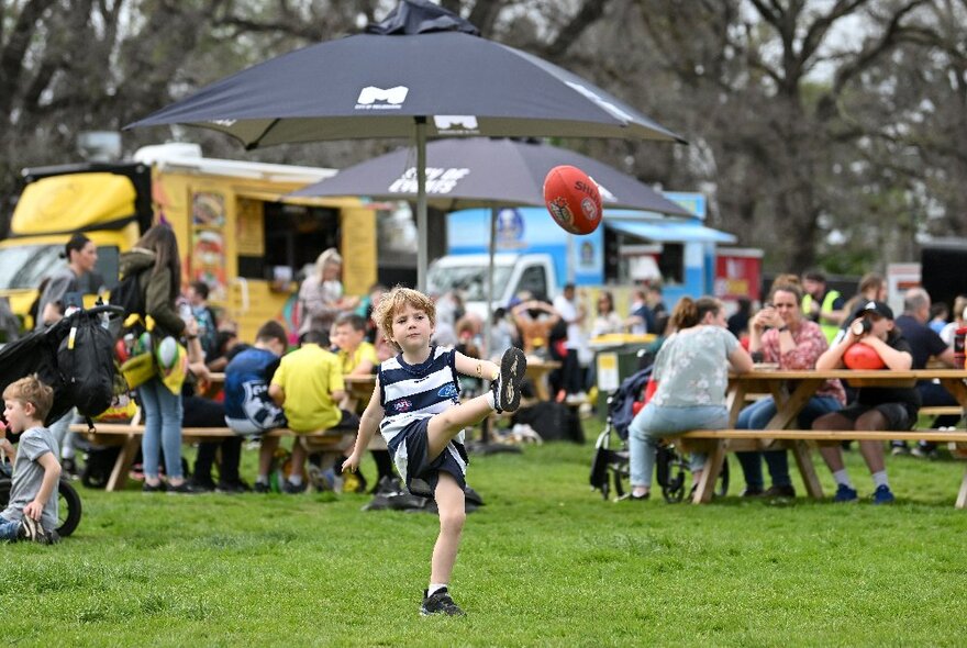 A young boy wearing his footy team's jumper, kicking a football outdoors, with crowds in the background, some seated at picnic tables in front of food trucks, enjoying the AFL Footy Festival in Yarra Park.