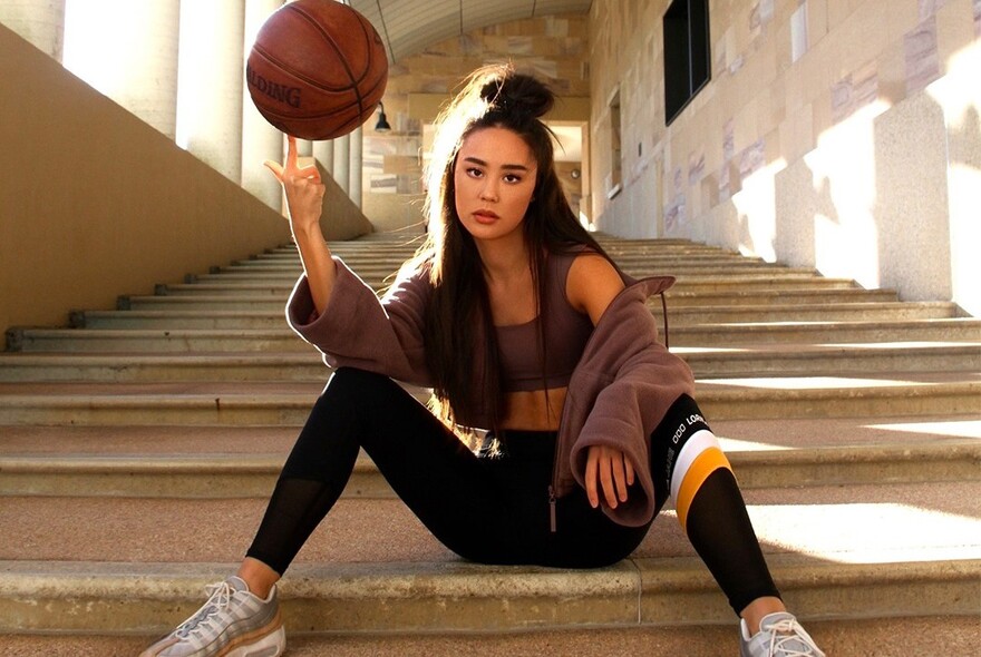 A model seated on stairs in active wear spinning a basketball on her finger. 
