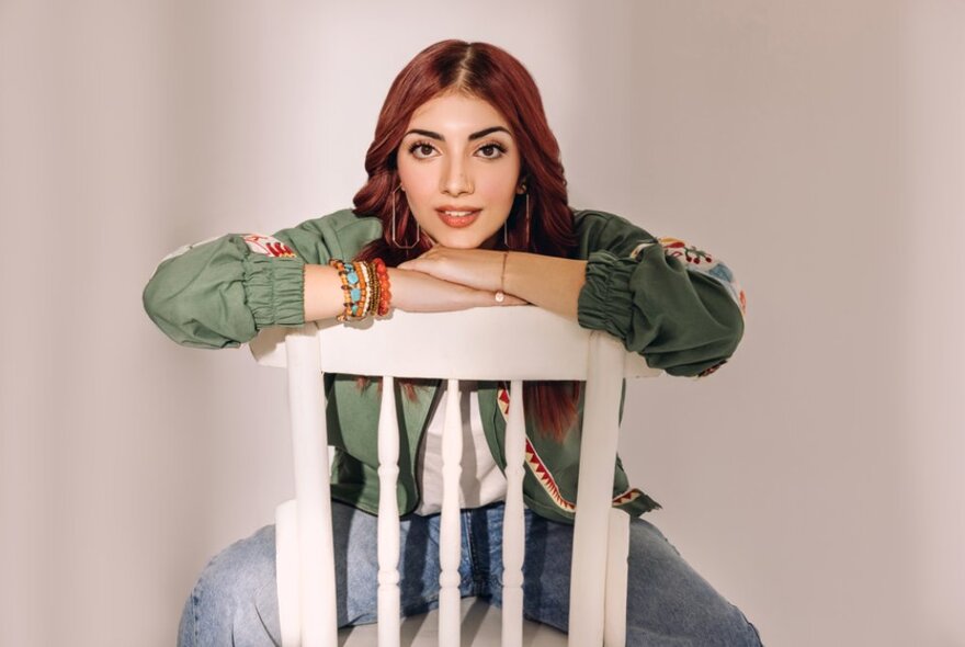 Female singer Annural Khalid, with long hair,  her arms resting under her chin straddling a white wooden chair.