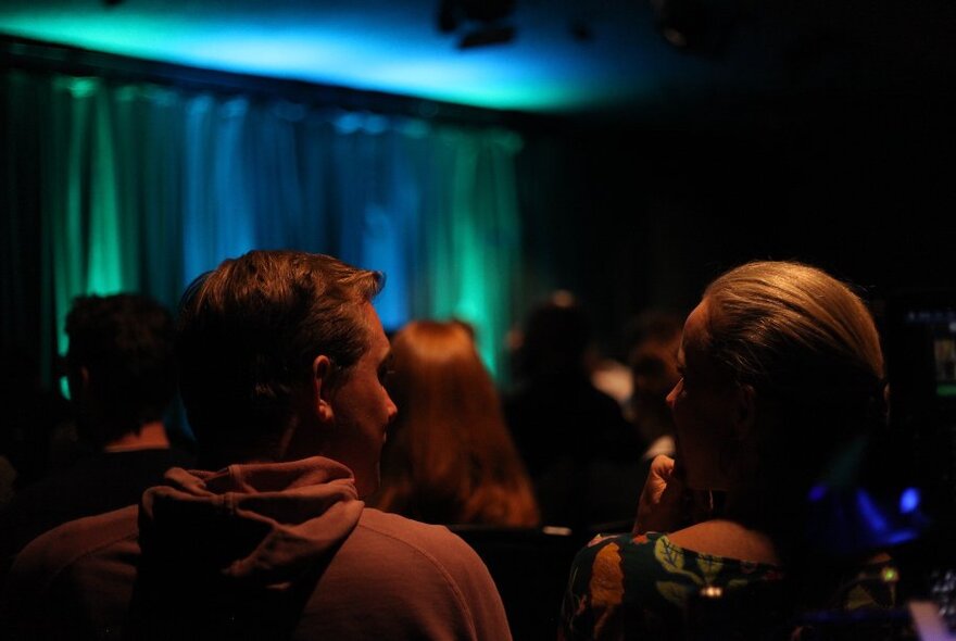 View of a small stage from the back rows of seated audience members, the back of heads silhouetted.