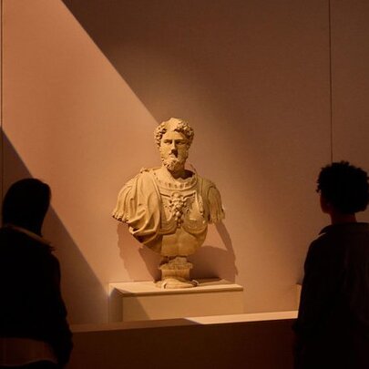 A marble bust of a Roman soldier lit up by a beam of light in a museum setting, people around it looking on.