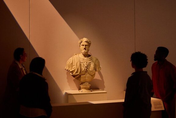 A marble bust of a Roman soldier lit up by a beam of light in a museum setting, people around it looking on.