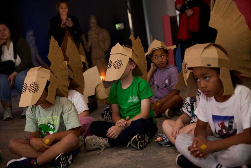 A group of seated children engaging in an interactive gladiator-themed workshop, the children are all wearing cardboard helmets.