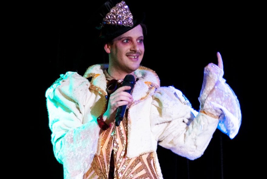 A smiling performer on stage wearing an elaborate velvet jacket and hat, holding a microphone.