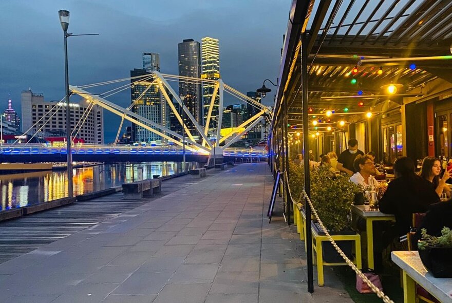 View of the exterior seating area of BangPop restaurant's outdoor terrace, with diners seated at tables, the wharf promenade, Yarra River and Melbourne's tall skyscrapers also visible; twilight.