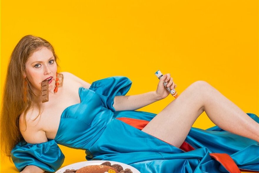 A woman reclining on the floor wearing a blue evening dress, stabbing her thigh with an epipen and eating chocolate at the same time.