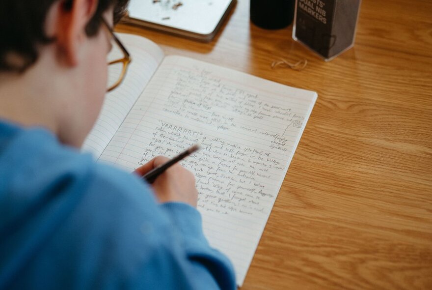 A young person seated at a table and writing longhand with a pen into a notebook.