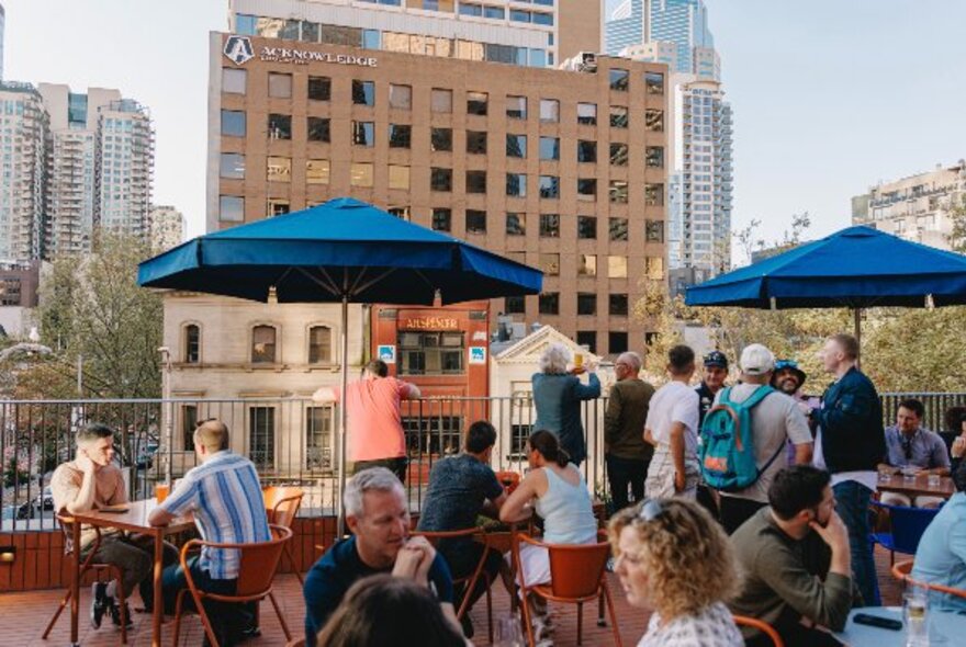 People seated at tables with umbrellas on an outdoor rooftop overlooking city buildings.