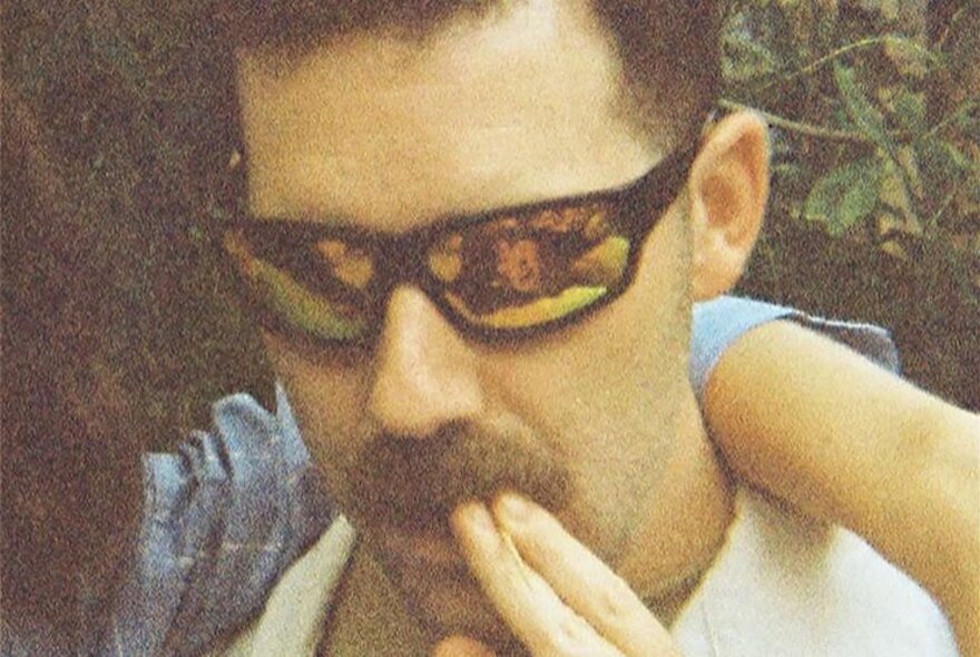 A grainy photo of a man with a moustache and mirrored sunglasses, an out-of-shot person's arm around him. 