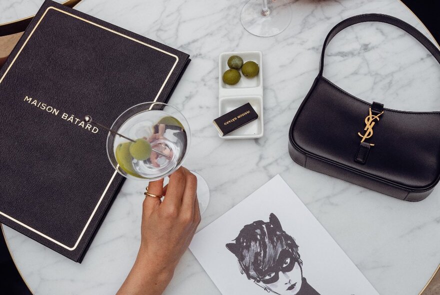 A white marble table with a menu for the restaurant Maison Batard, a hand holding a martini, a drawing of Catwoman, a YSL handbag and some olives.